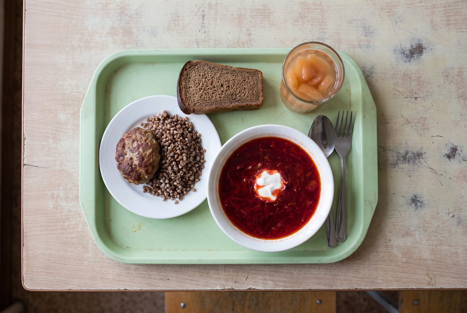 A Russian school canteen tray from above: borscht with soured cream, cutlet with buckwheat, rye bread and a glass of compote