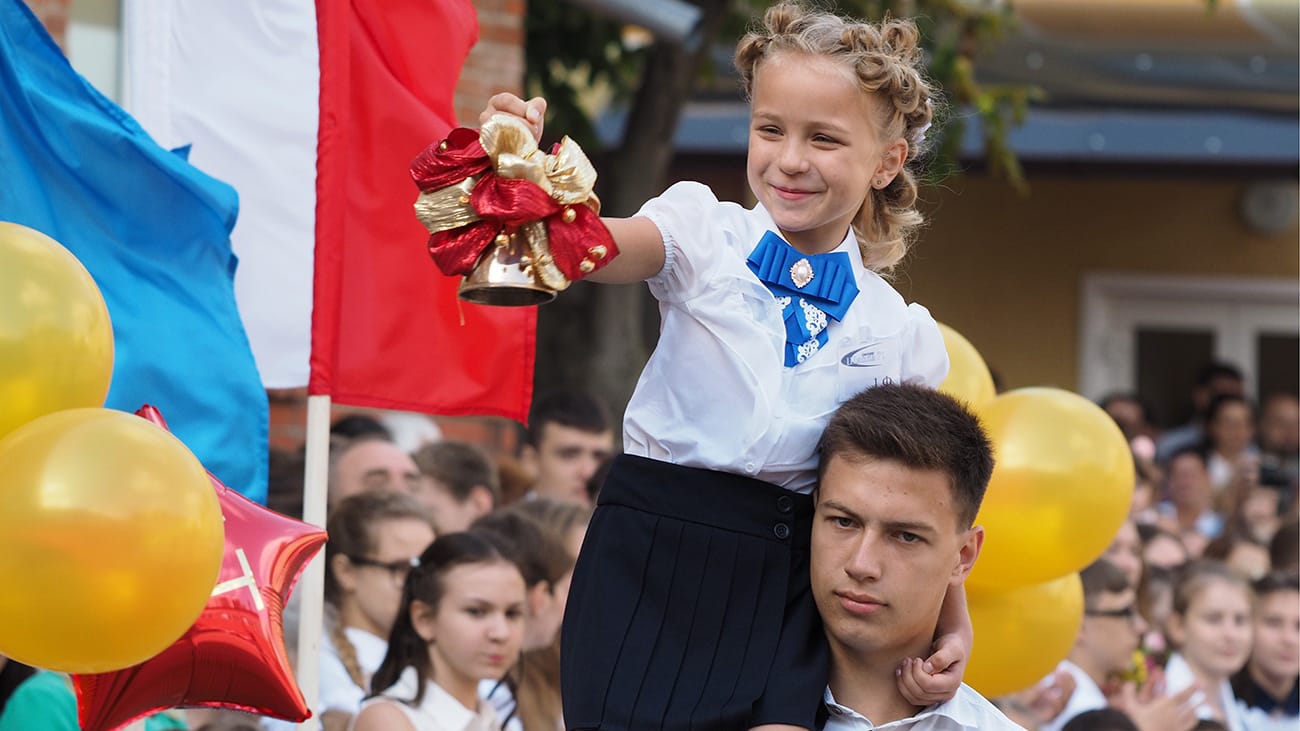 A young Russian pupil holding a decorated handbell at a Knowledge Day ceremony, surrounded by classmates and balloons